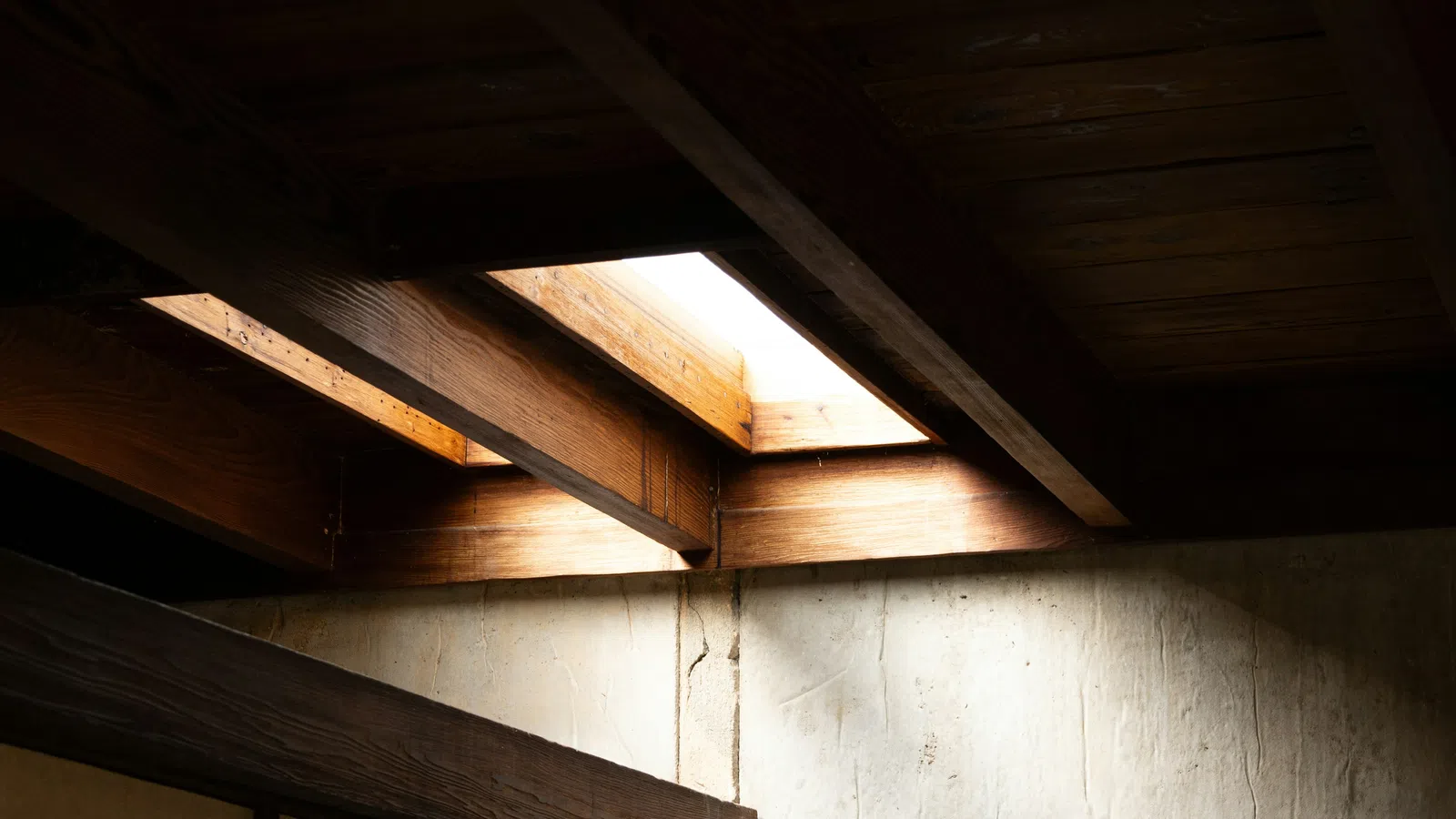 Natural light shines through wooden ceiling beams onto a textured concrete wall in a minimalist interior.