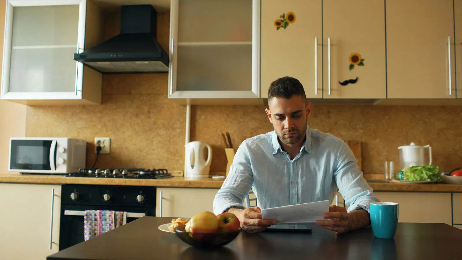 Man reading documents at kitchen table with fruit bowl and coffee mug, cabinets with sunflower stickers in background
