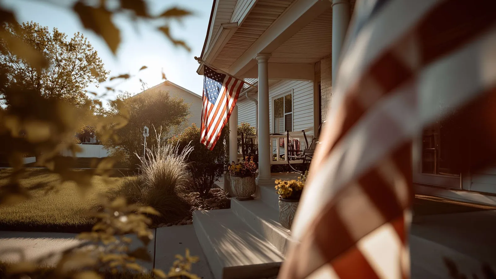 American flag hanging on a suburban house porch in warm sunlight with plants and lawn in background