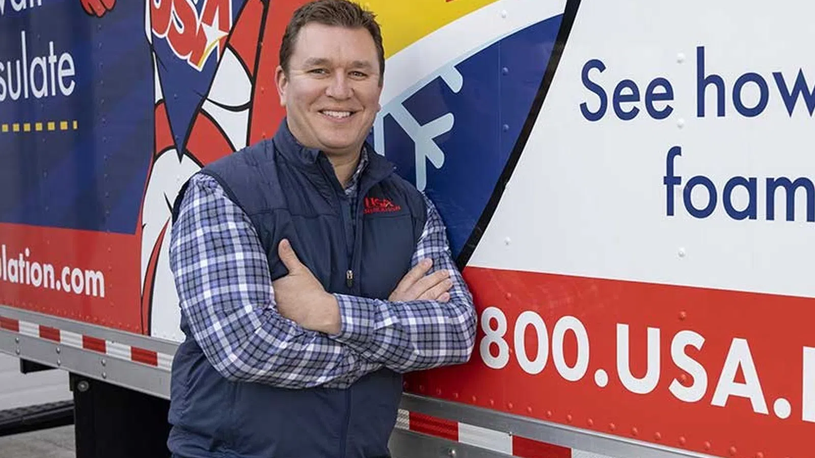 Smiling man in a plaid shirt and vest stands with arms crossed by a colorful insulation service truck.