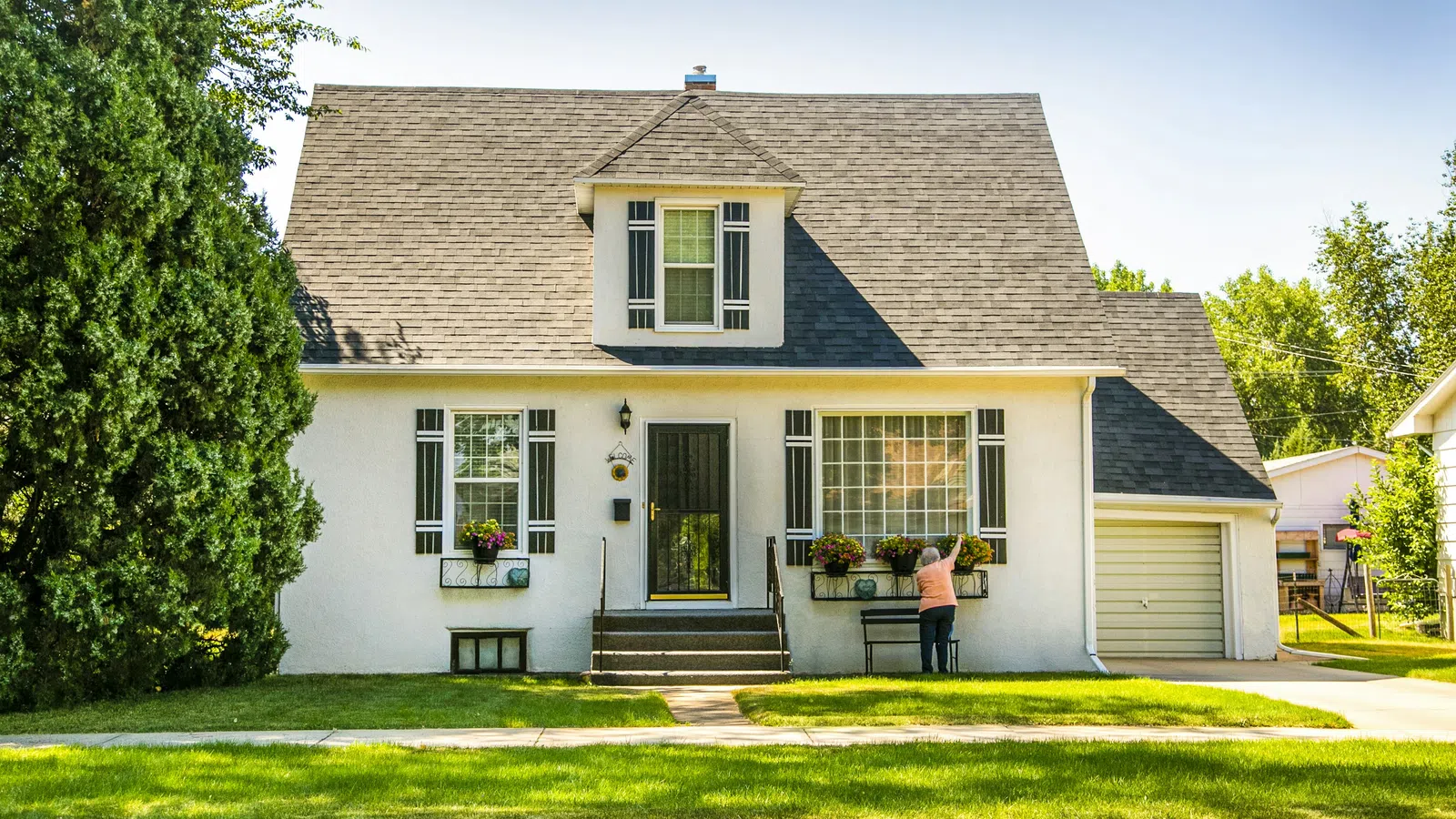Cozy white house with dark shutters, green lawn, and person tending to window flower boxes on a sunny day