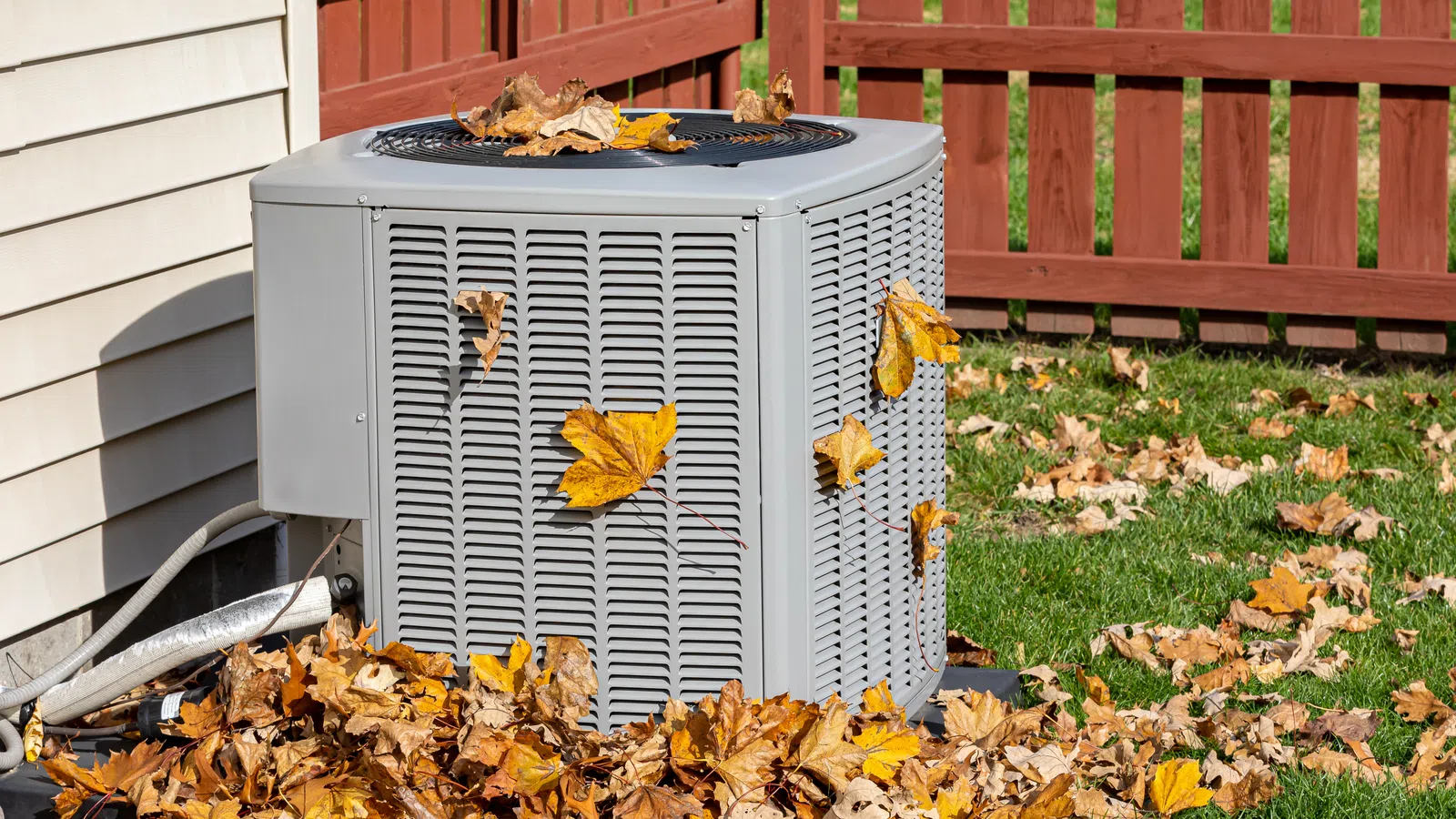 An air conditioning unit covered in autumn leaves near a wooden fence and grassy area.