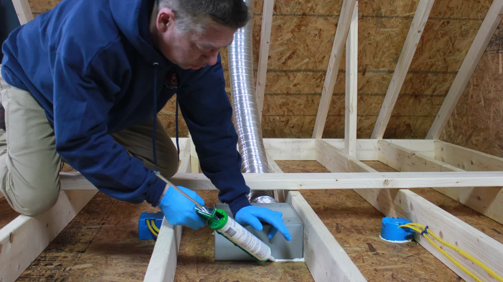 Worker in blue hoodie and gloves applying sealant to ventilation duct in attic framed with wood beams.