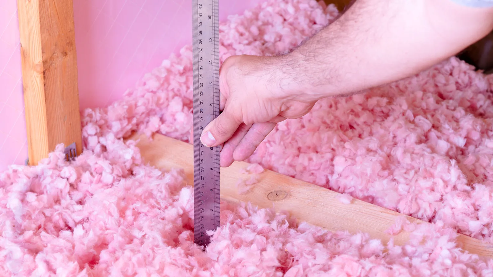 Hand measuring depth of pink fiberglass insulation with metal ruler inside attic wooden frame.