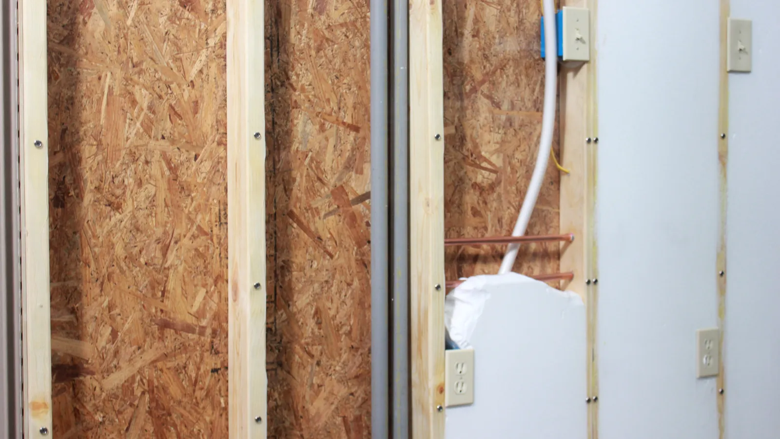Unfinished wall with exposed wood studs, OSB panels, electrical outlets, and pipes in a home under construction or renovation.