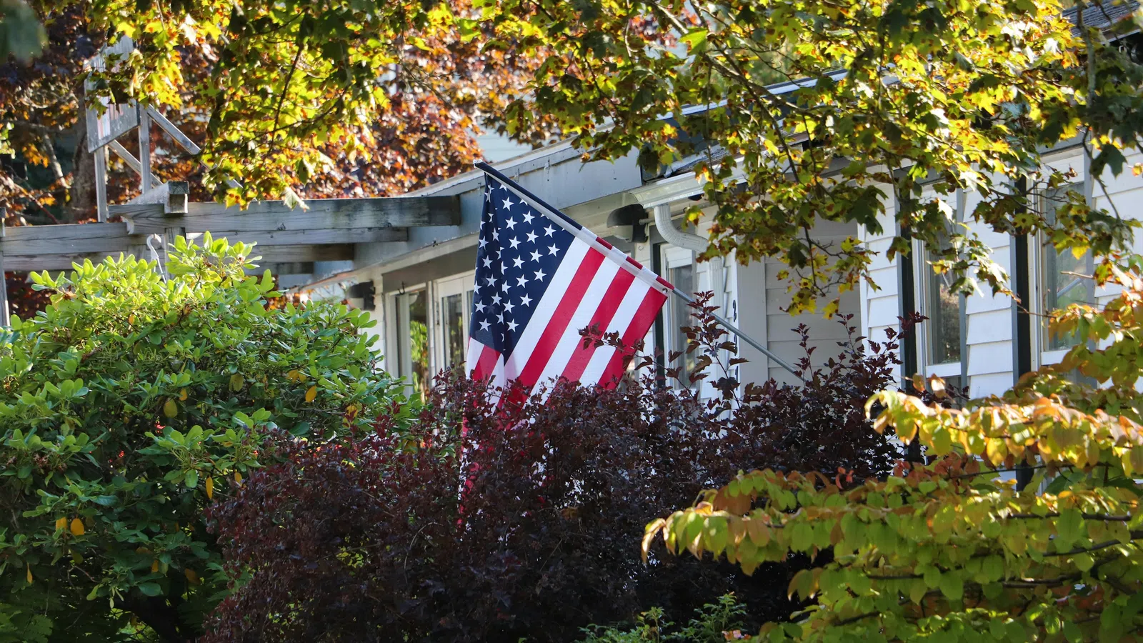 American flag displayed on a house porch surrounded by lush green and autumnal foliage in daylight.