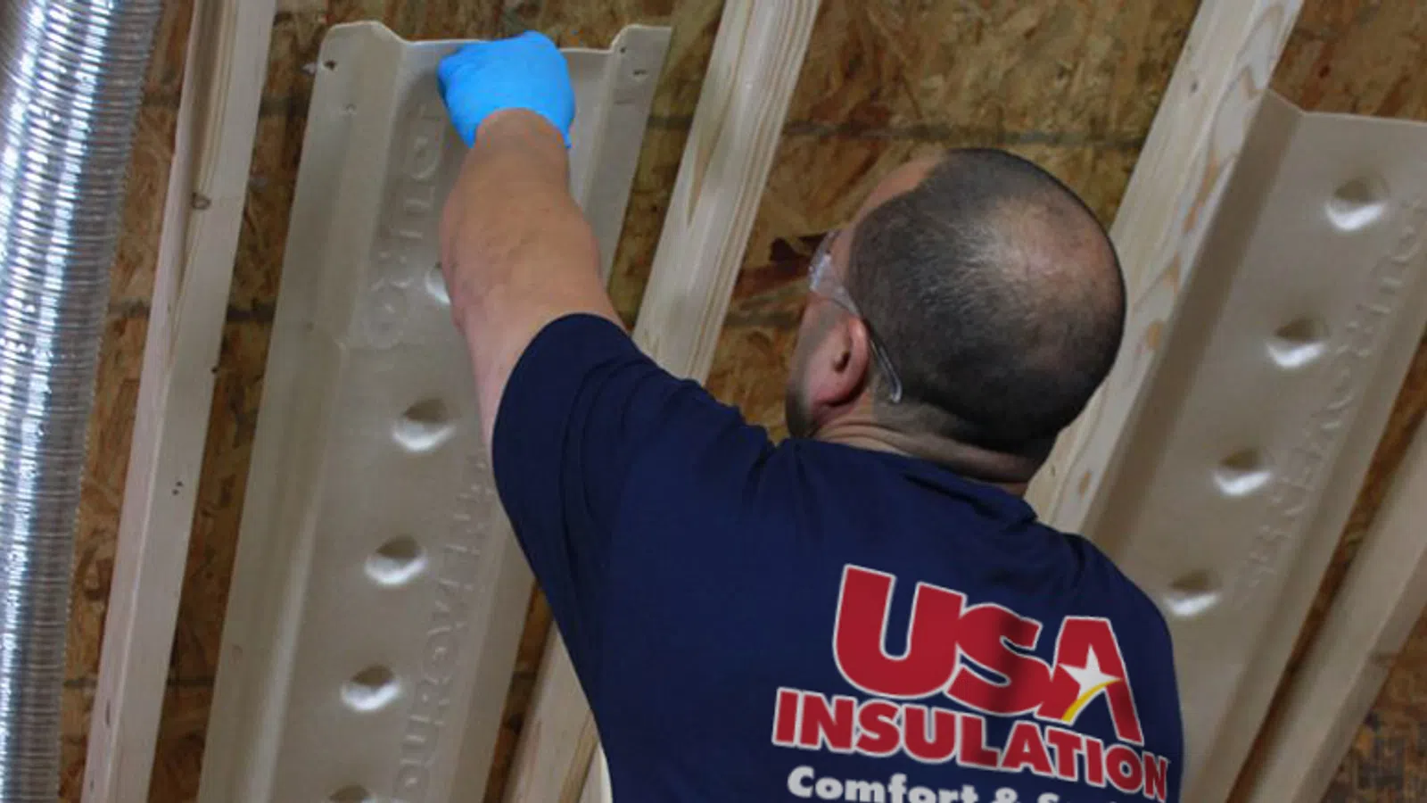 Worker wearing blue gloves installing insulation panels on an attic ceiling with exposed wooden beams.