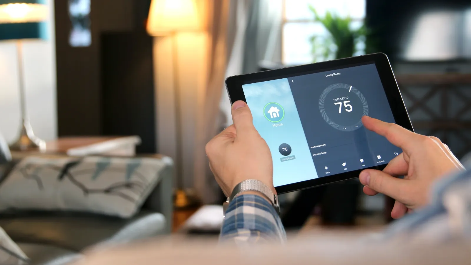 Person adjusting smart thermostat settings on a tablet in a cozy living room with warm lighting.