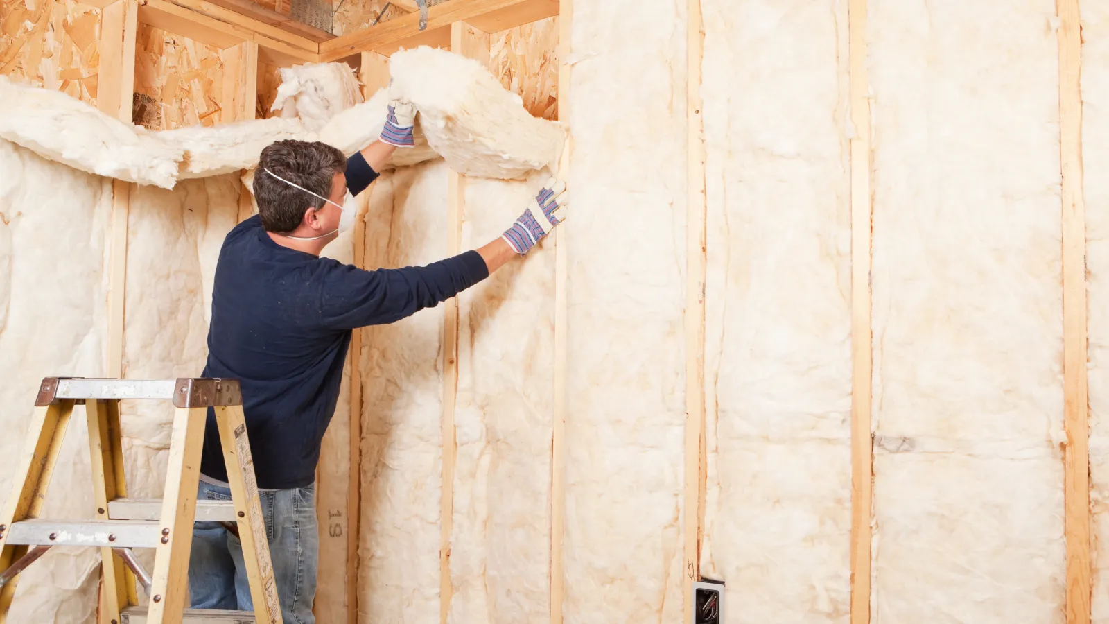 Worker installs fiberglass insulation on wooden frame wall inside a house under construction.