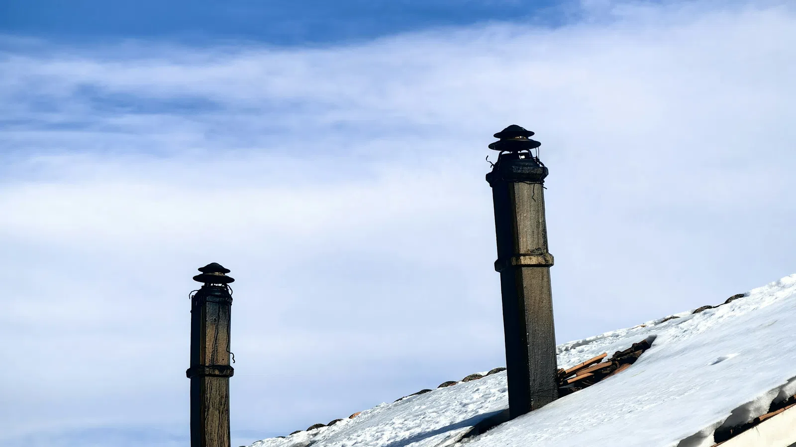 Snow-covered brick house roof with two black chimneys under blue sky with clouds.