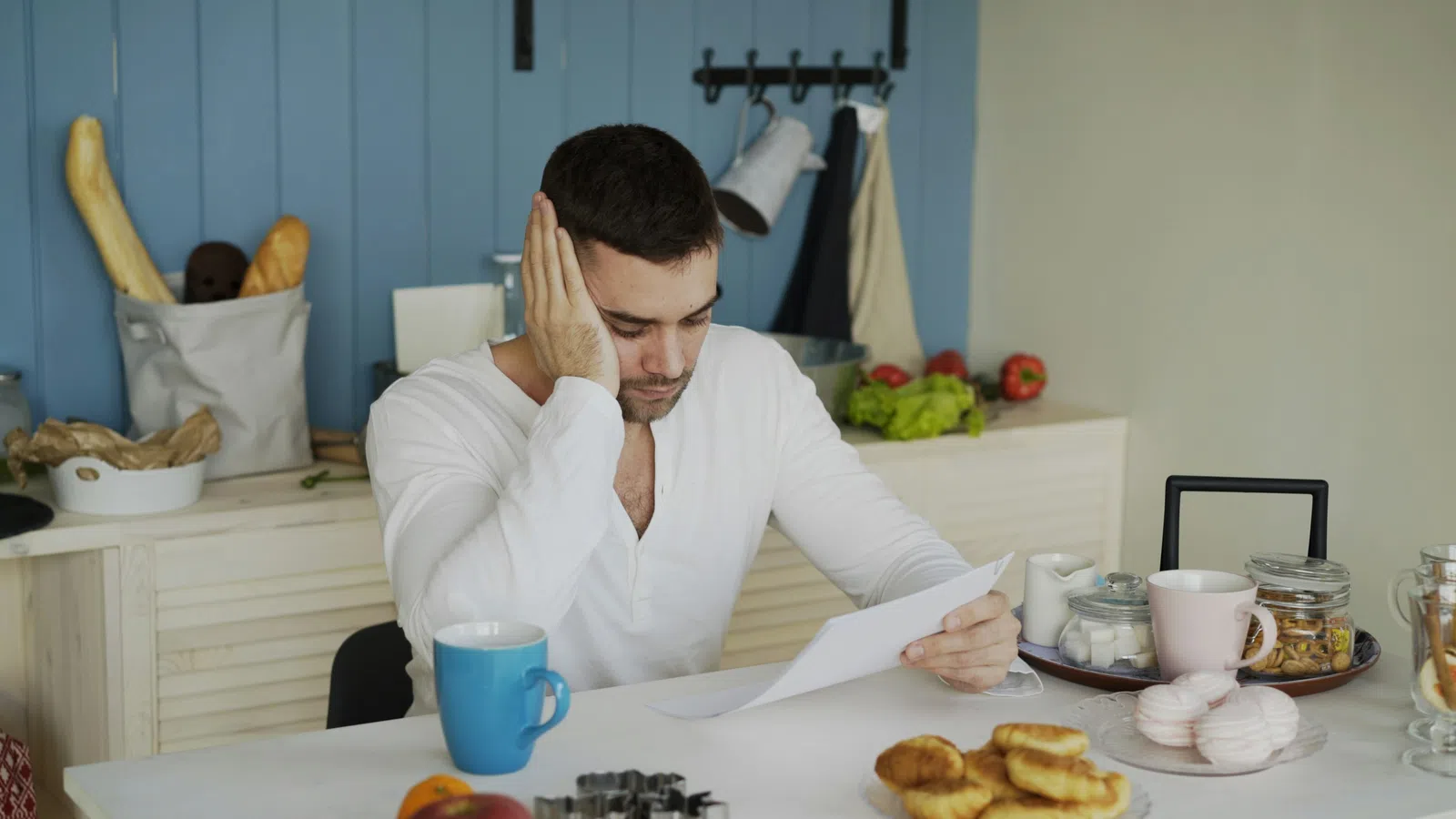 Man in white shirt reading documents at kitchen table with pastries, fruit, jars, and bread in the background