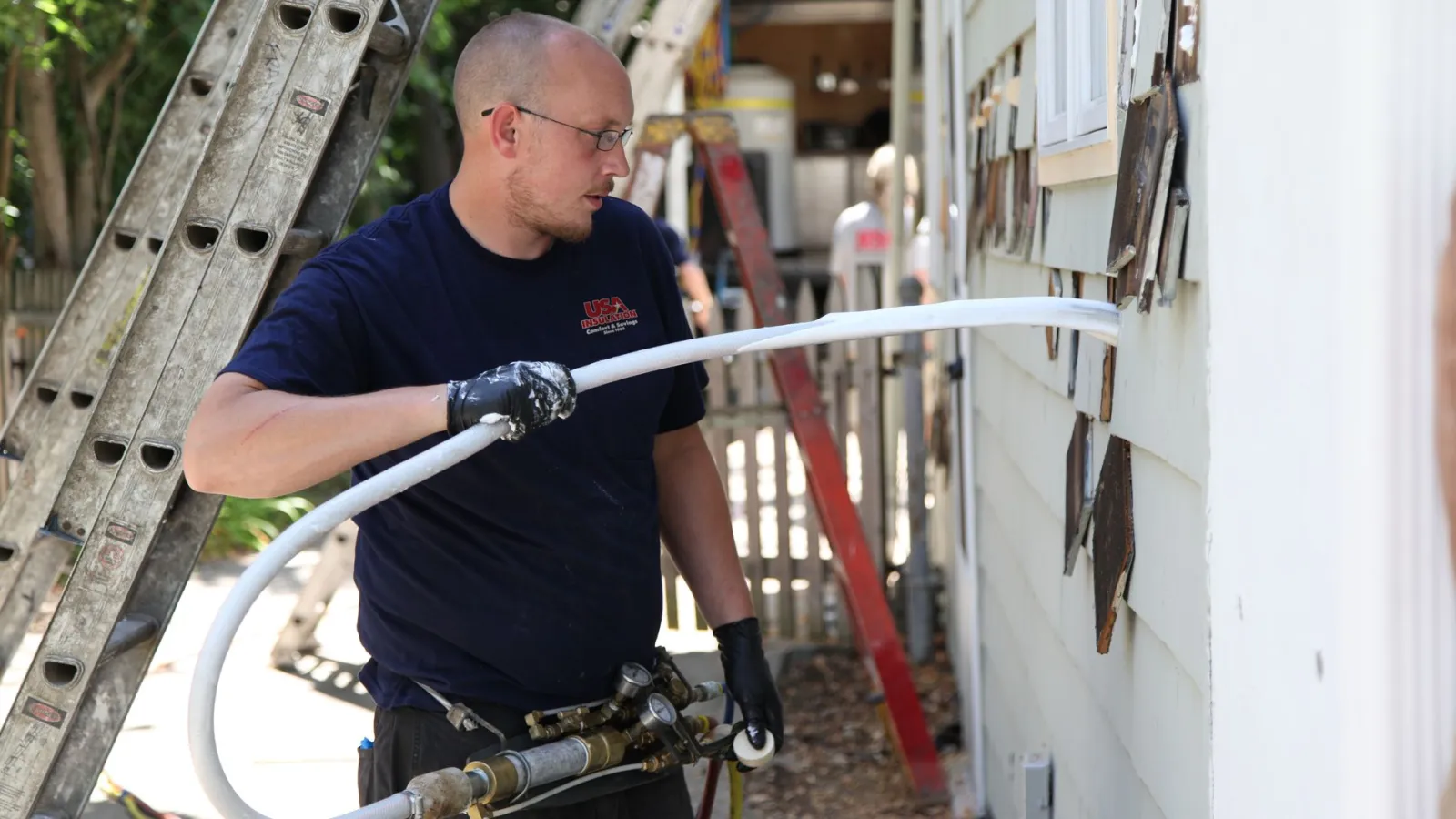Technician wearing gloves feeds a white cable through an exterior house wall for installation or repair work.
