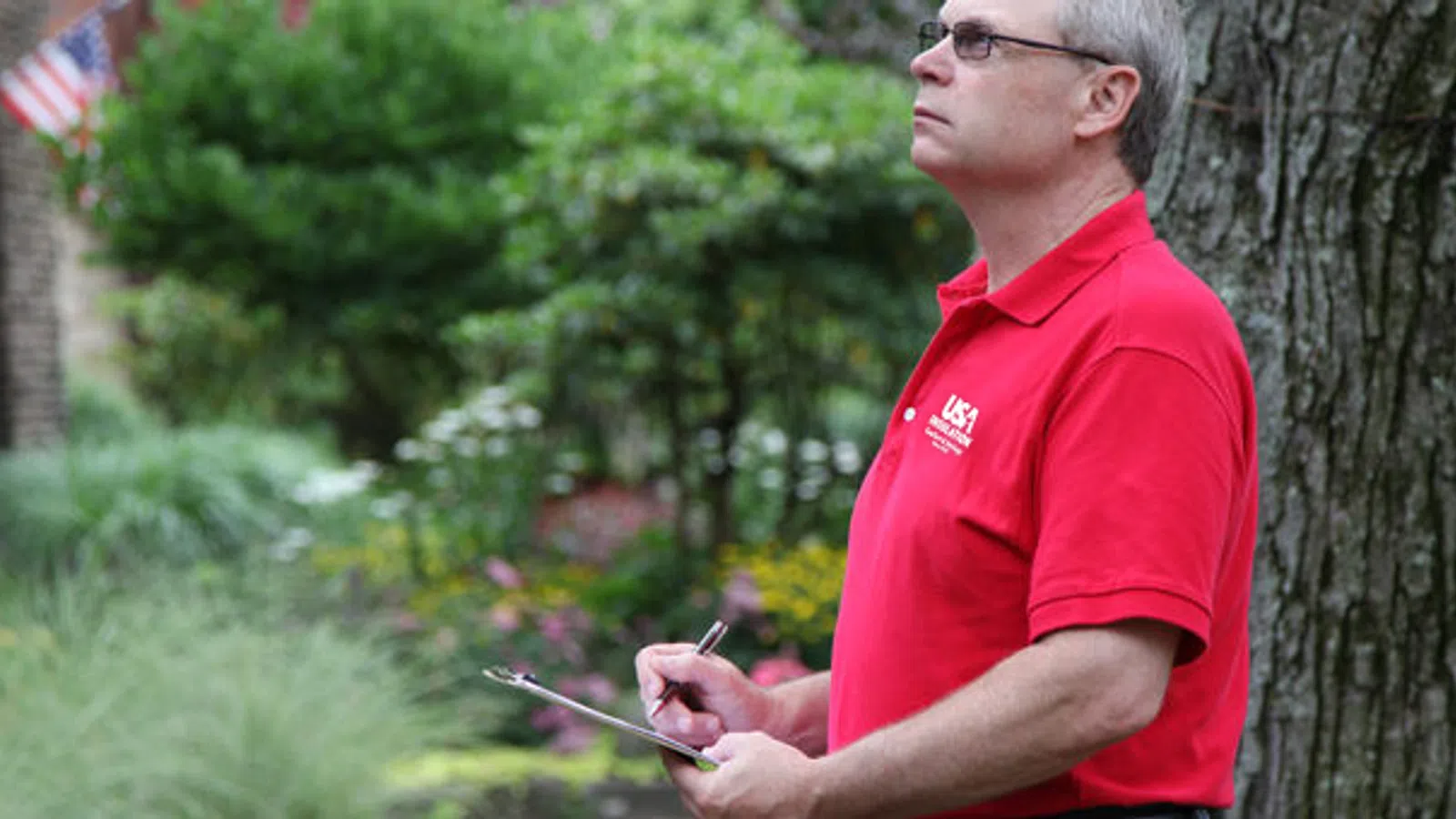 Man in red shirt holding clipboard and pen outdoors near tree writing or inspecting in a residential area