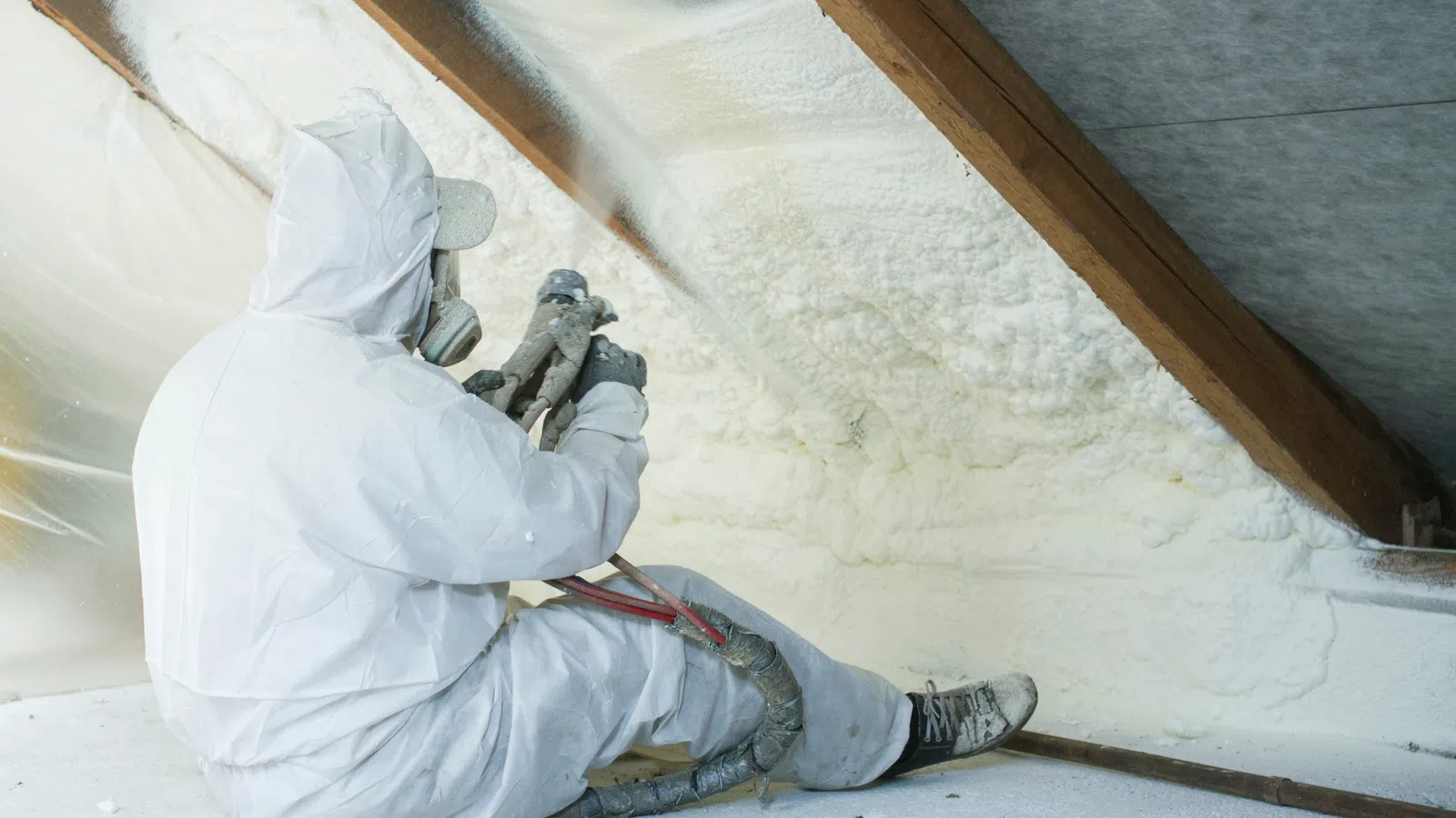 A worker in protective gear applies spray foam insulation in an attic space.