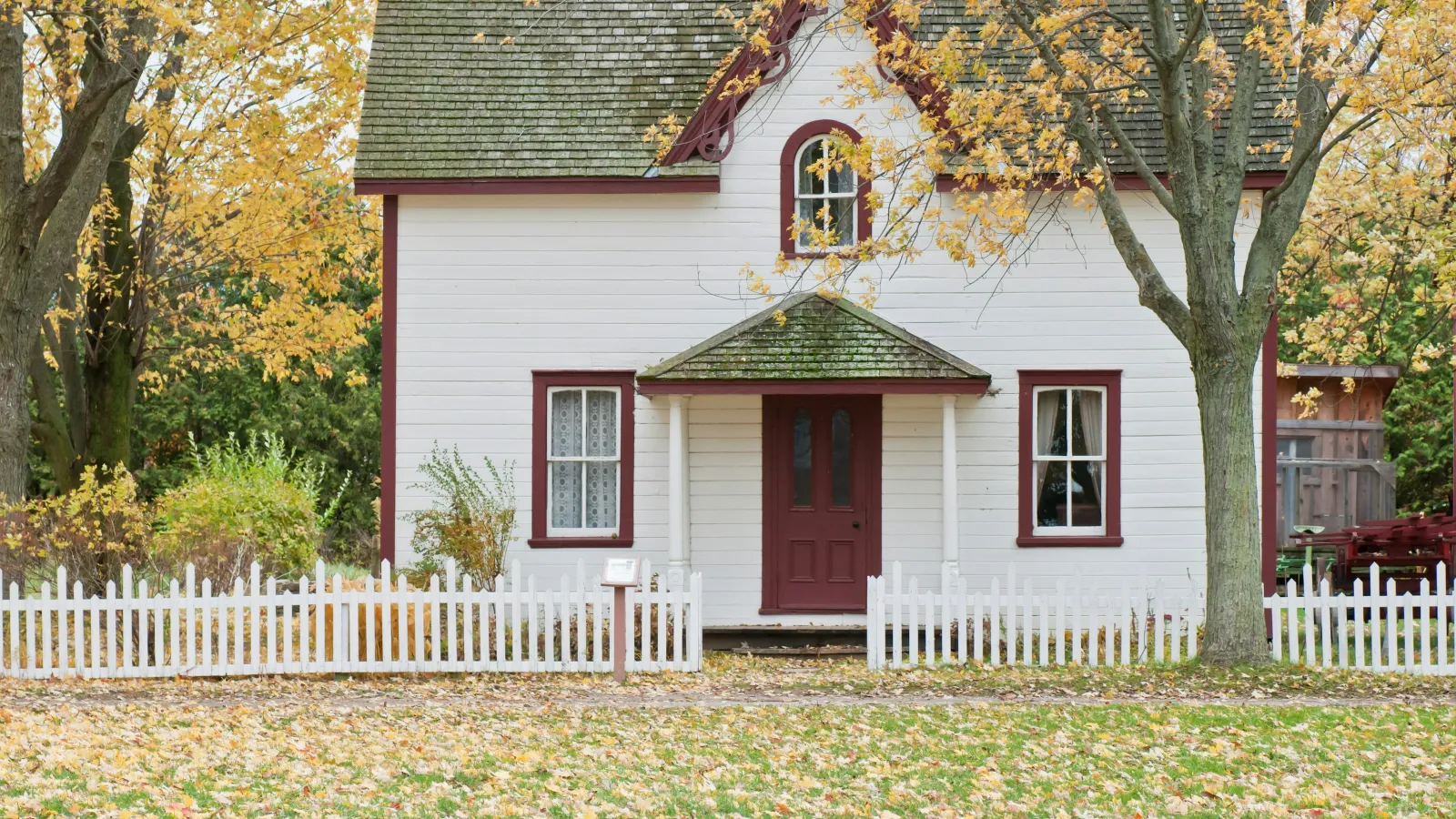 White house with red trim surrounded by fall trees and a white picket fence with autumn leaves on the ground