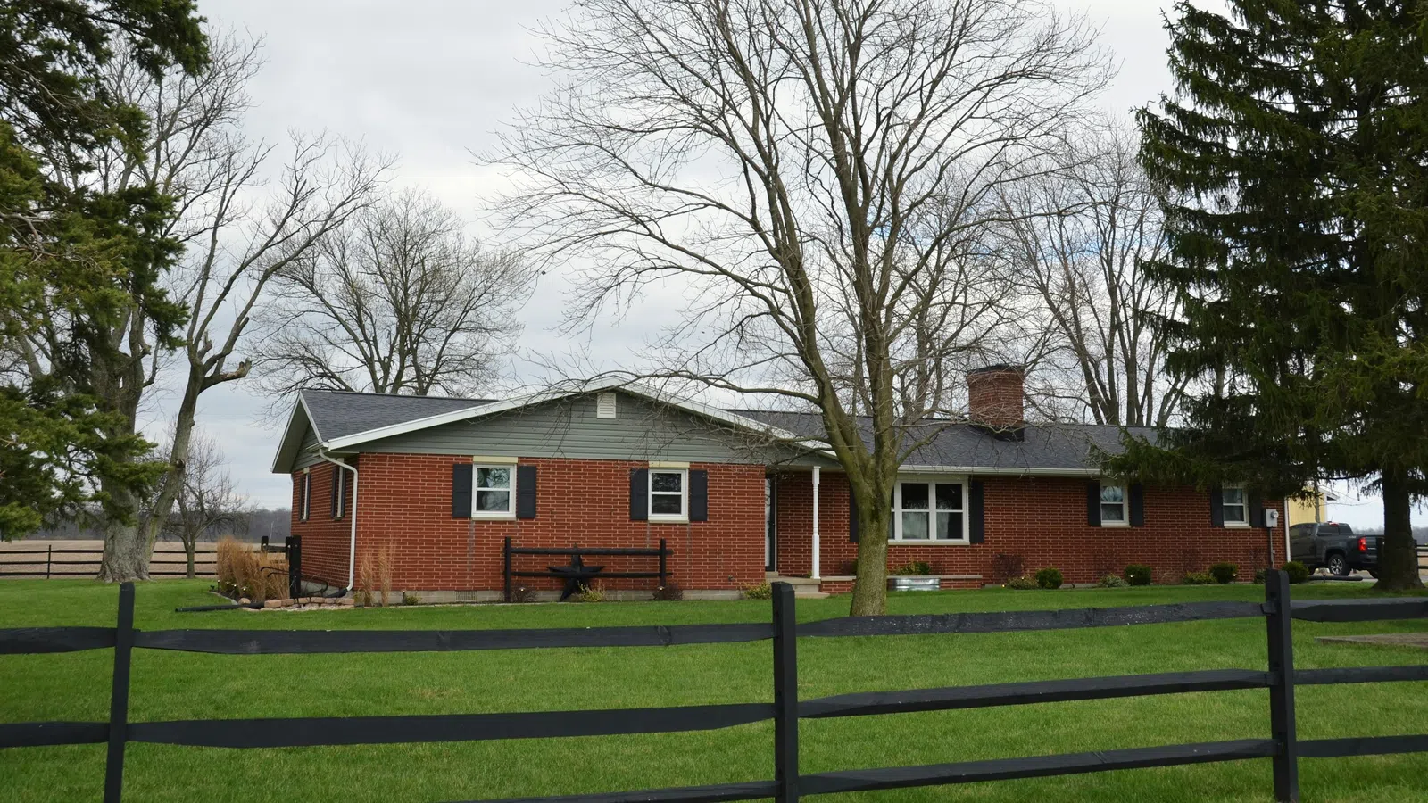 One-story red brick house with black shutters behind black wooden fence on green lawn under cloudy sky