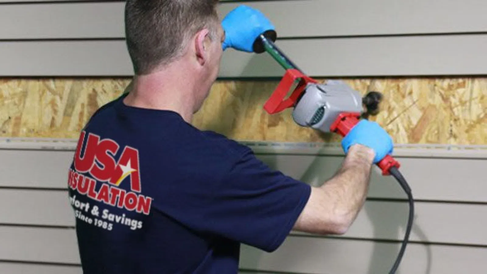 Worker wearing blue gloves using power tool to install beige siding on a building exterior.