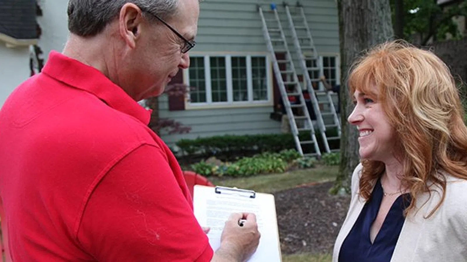 Man in red shirt taking notes on clipboard while talking to smiling woman outdoors near house