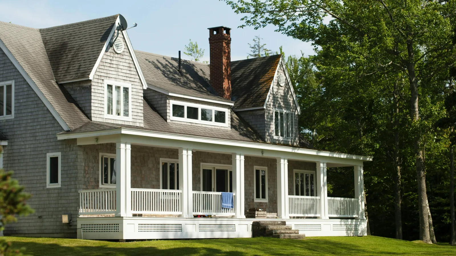 Spacious gray shingle house with white porch railing and lush green lawn surrounded by trees on sunny day