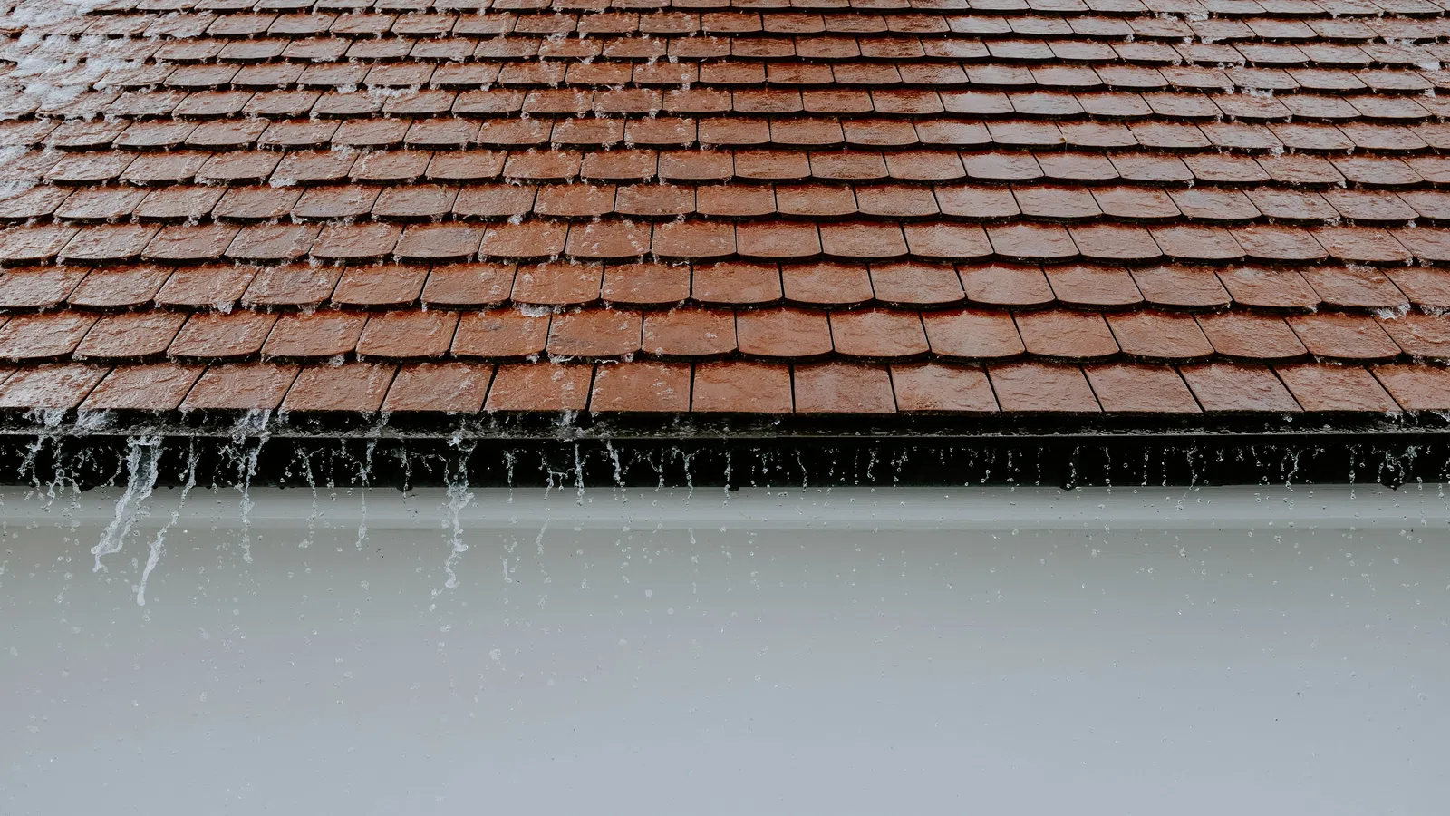Red clay roof tiles with water dripping off the edge during rain on a gray sky background.