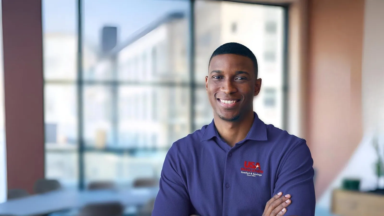 Smiling man in blue USA heating company shirt standing with arms crossed in modern office space.