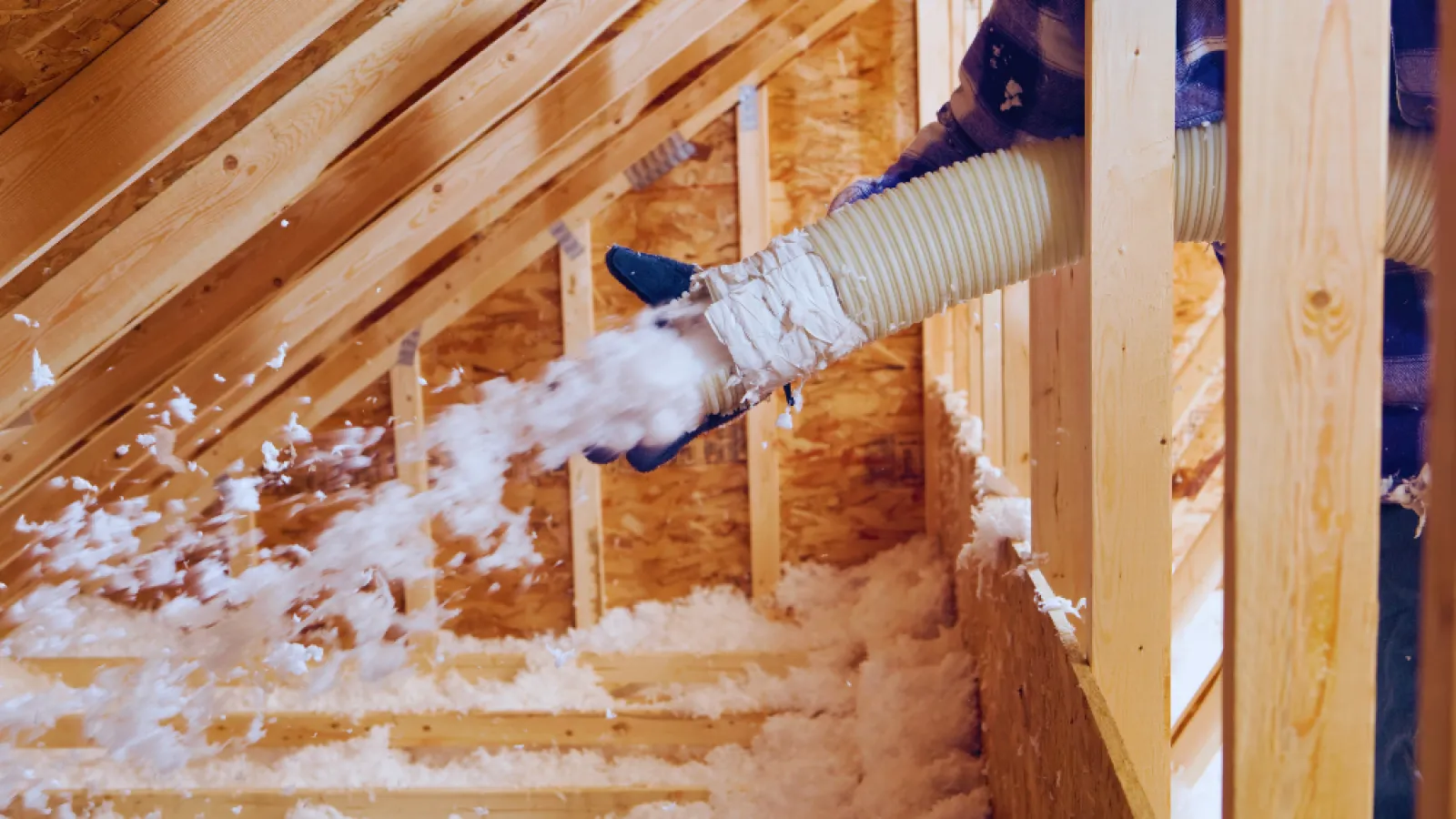 Worker installing loose-fill insulation in an attic using a hose to spread material evenly.
