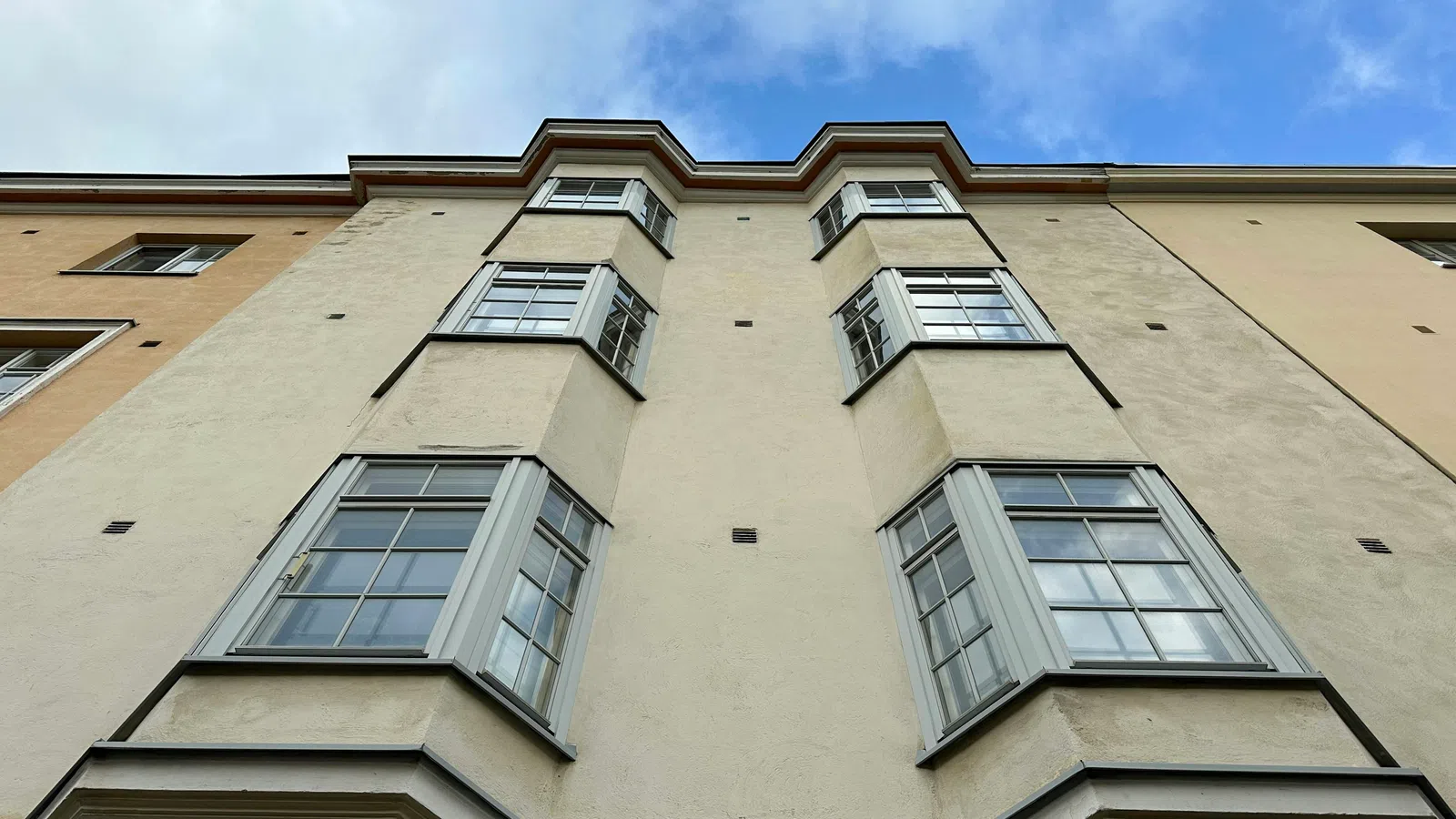 Low angle view of beige building facade with bay windows under a blue sky with clouds.