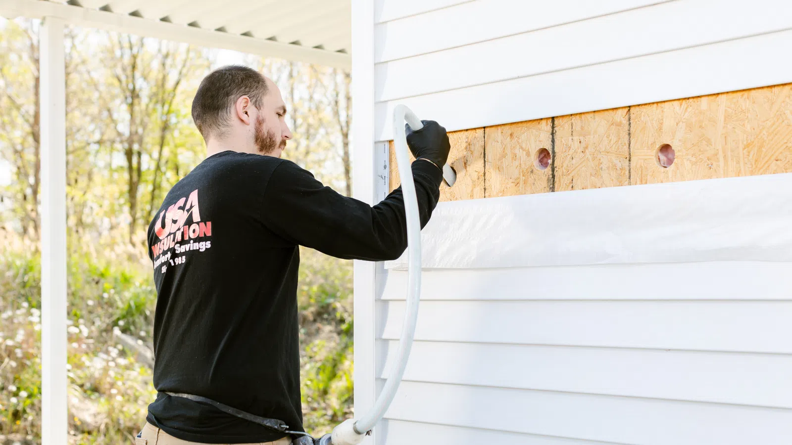 A handyman applying insulation to a home's exterior wall with a spray nozzle, showcasing renovation work.