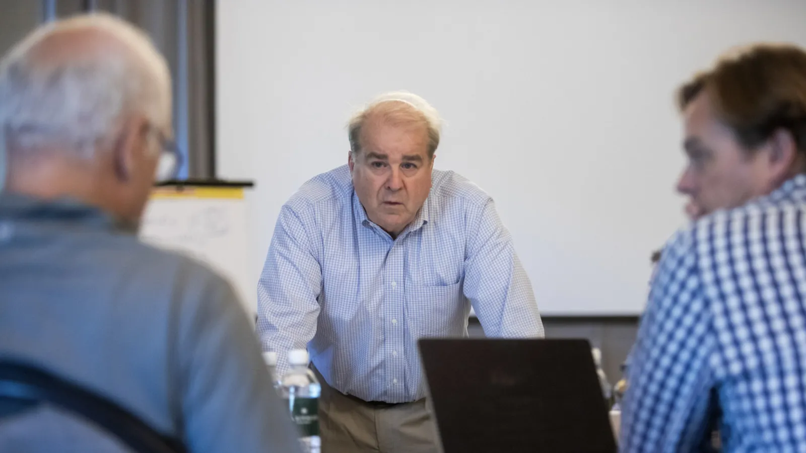Senior man in blue checkered shirt leading a meeting with two attendees focused on discussion in a conference room.