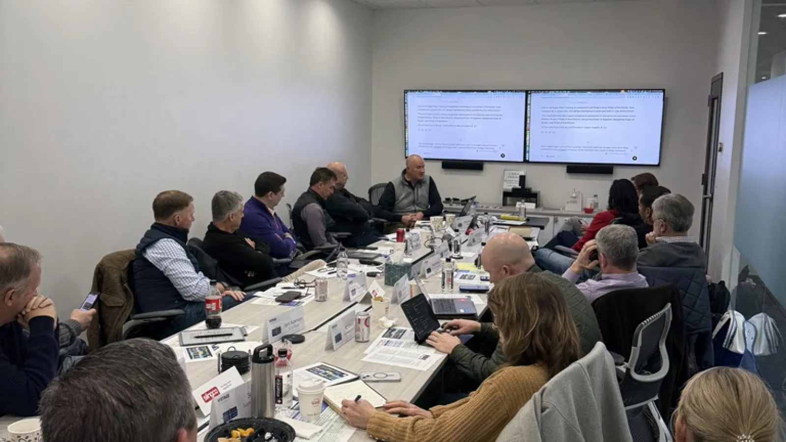 Business professionals engaged in a meeting around a long conference table with screens displaying presentation content.