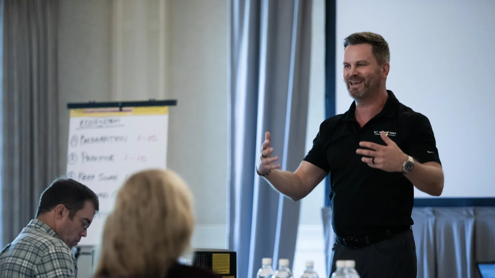 Man in black shirt giving presentation to seated attendees in a conference room with flip chart and water bottles.