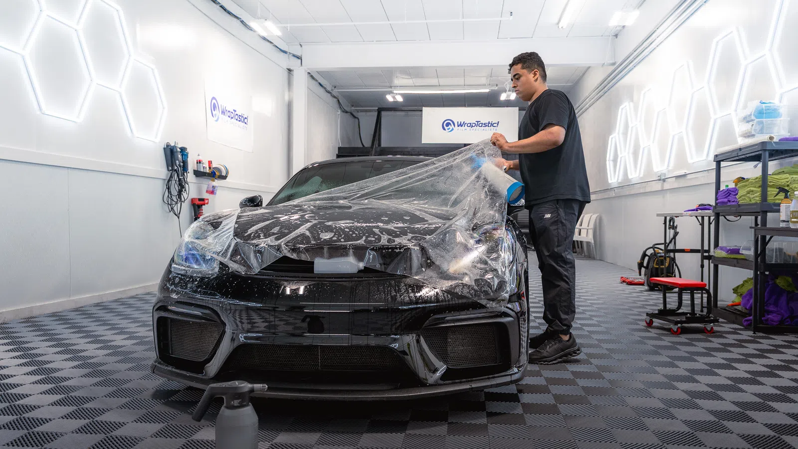 Technician applies protective film to black sports car's hood inside a modern auto detailing studio.