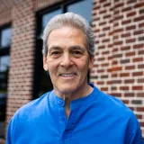 Smiling older man with gray hair wearing a blue shirt standing outside of Restore in 24 near a brick wall and windows.