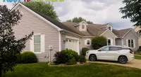 Suburban house with beige siding, green lawn, white SUV in driveway, and cloudy sky with Roof It Forward logo.
