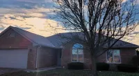 Brick house with large garage and leafless tree in front under a cloudy sky at sunset.