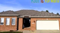 Brick house with gray shingled roof, white garage door, and a dog sitting at the front steps under clear blue sky.
