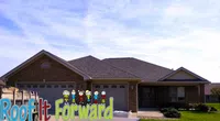 Single-story brick house with dark shingled roof under blue sky with Roof It Forward text and cartoon figures.