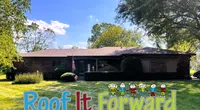 Single-story brick house with a manicured lawn under a clear blue sky, featuring Roof It Forward logo.