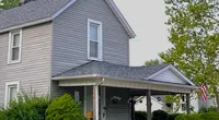 Gray two-story house with new roof, front porch, green shrubs, and white flowers under bright sky with Roof It Forward logo.