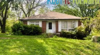Brick house with beige roof surrounded by green trees and lawn under a clear sky with Roof It Forward logo.