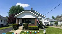 Single-story brick and white house with a porch, American flag, and Roof It Forward sign on the lawn under a clear blue sky.