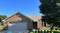 Brick house with a double garage and a Roof It Forward sign with colorful letters and children figures on the lawn.