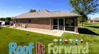 Brick house with a shingled roof, green lawn, and Roof It Forward logo with cartoon children outdoors.