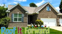 Brick house with a gray roof and a green lawn displaying a colorful Roof It Forward sign with cartoon children.