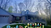 Suburban house with a porch and colorful Roof It Forward sign on the lawn surrounded by trees in early evening light.