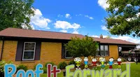 Brick house with a new roof under a blue sky, featuring the Roof It Forward logo and cartoon children on the lawn.