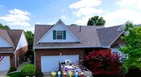 Suburban brick house with white garage filled with roofing debris under clear sky and Roof It Forward logo.