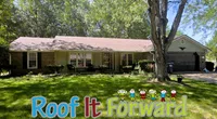 Single-story brick house with large tree and green lawn under a clear blue sky with Roof It Forward text.