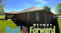 Single-story brick house with green lawn under clear blue sky and Roof It Forward logo with playful kid illustrations.