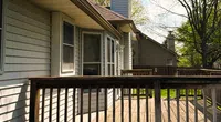Sunny wooden deck attached to a house with beige siding and trees in the background on a clear day.